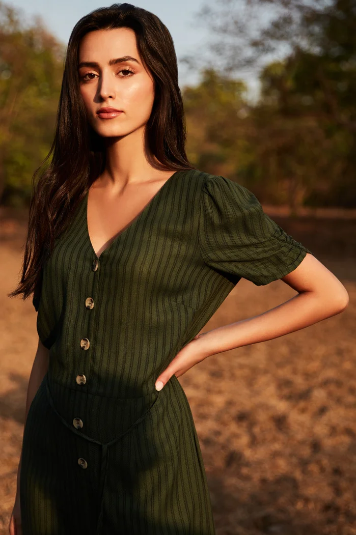 A woman with long, dark hair wearing a green striped dress with buttons, posing confidently in a natural outdoor setting.