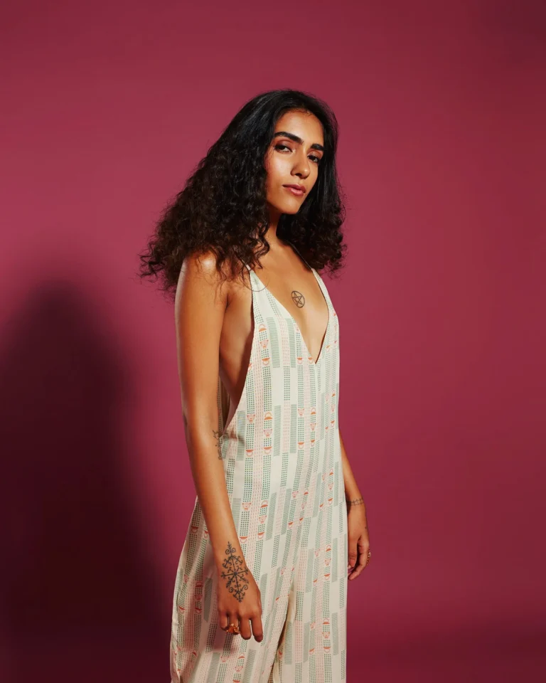 A woman with curly hair wearing a patterned jumpsuit, standing confidently against a pink background.