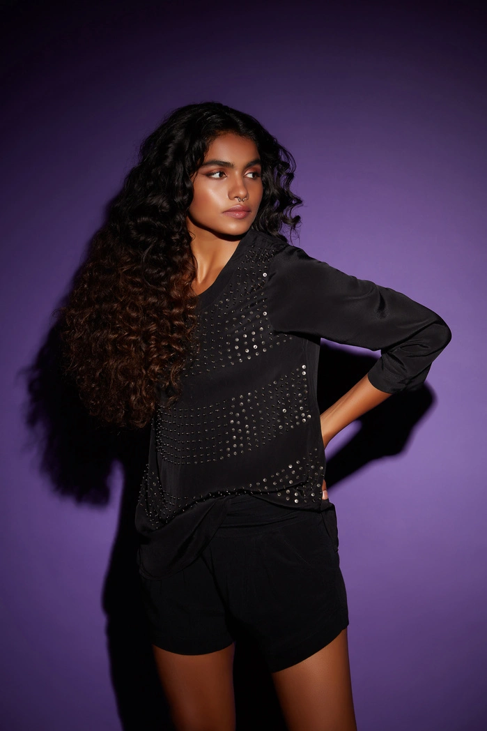 A woman with long curly hair wearing a black top with embellishments, posing against a purple background.