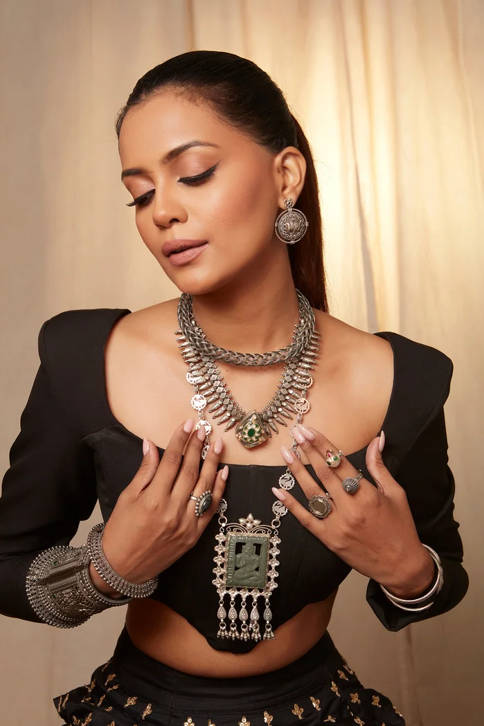 A woman with long hair wearing a black outfit adorned with intricate silver jewelry, posing gracefully with her hands on her jewelry.