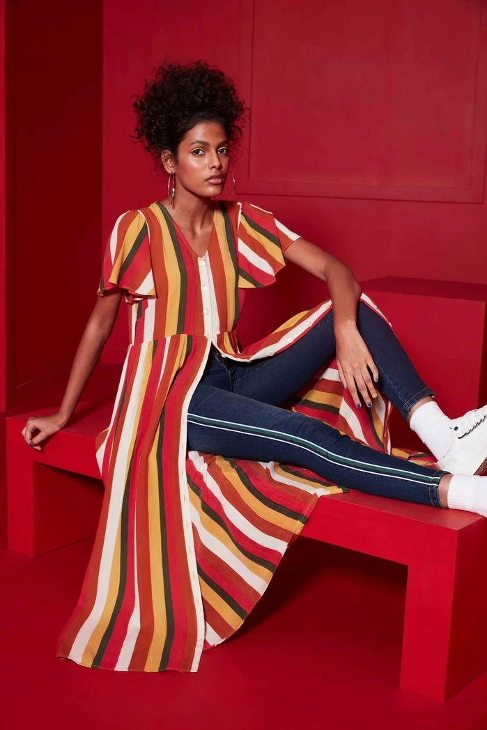 A woman with curly hair wearing a striped dress over jeans, sitting on a red bench.