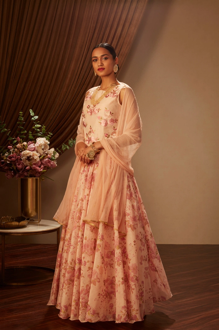 A woman in a pink floral lehenga, standing elegantly with a floral arrangement nearby.
