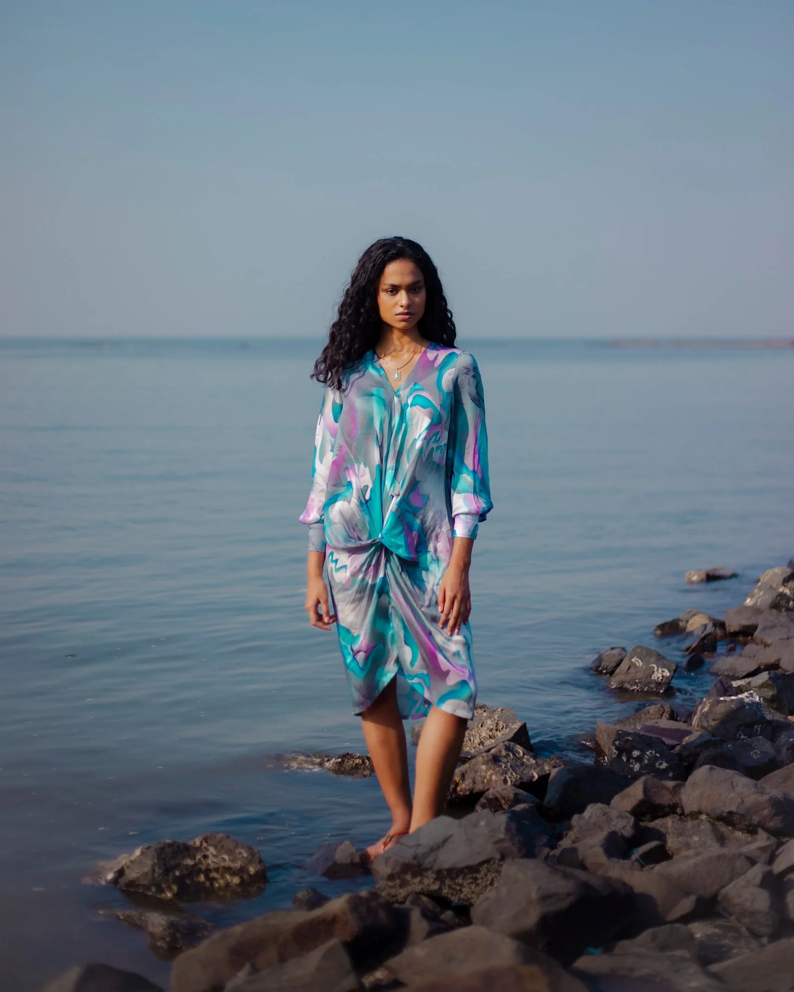 A woman standing on rocky shore by the water, wearing a colorful dress.