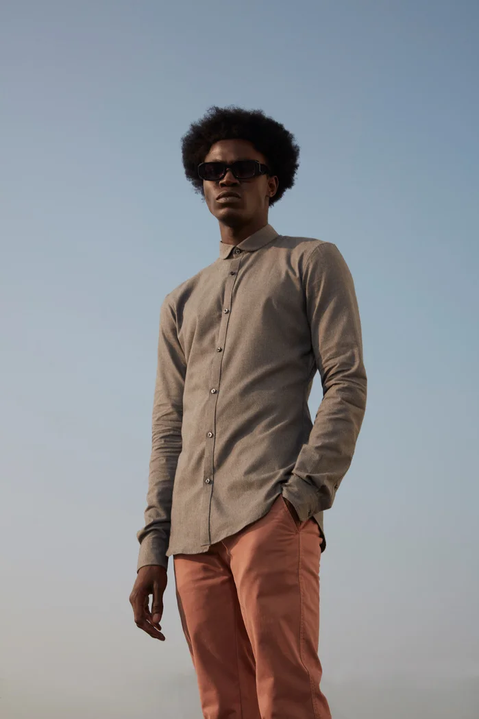 A man with an afro hairstyle wearing a gray button-up shirt and pink pants, standing confidently against a clear sky.