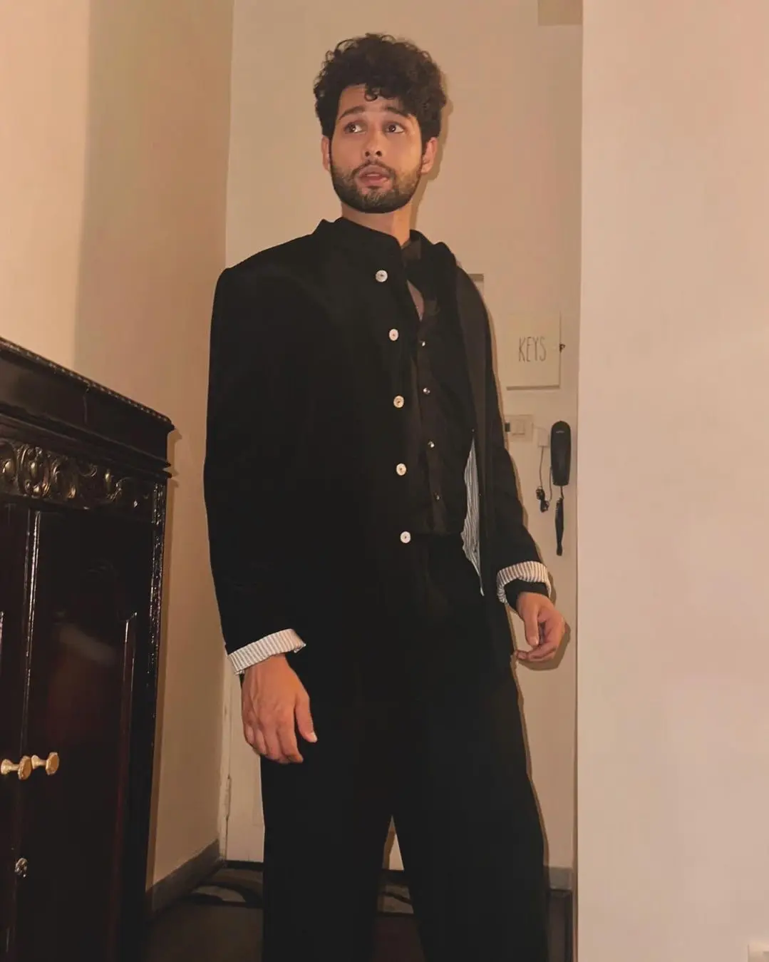 Siddhant Chaturvedi wearing a black outfit with a stylish jacket, standing in a hallway with a thoughtful expression.