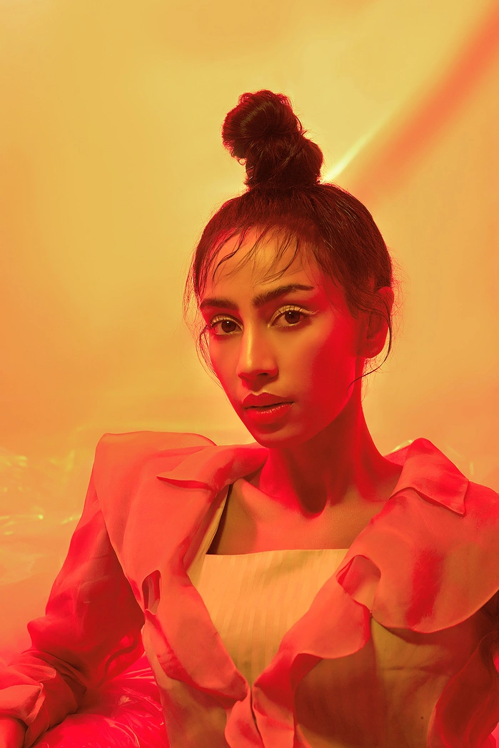 A woman with a top bun hairstyle, wearing a ruffled pink blouse, against a warm yellow-orange background.